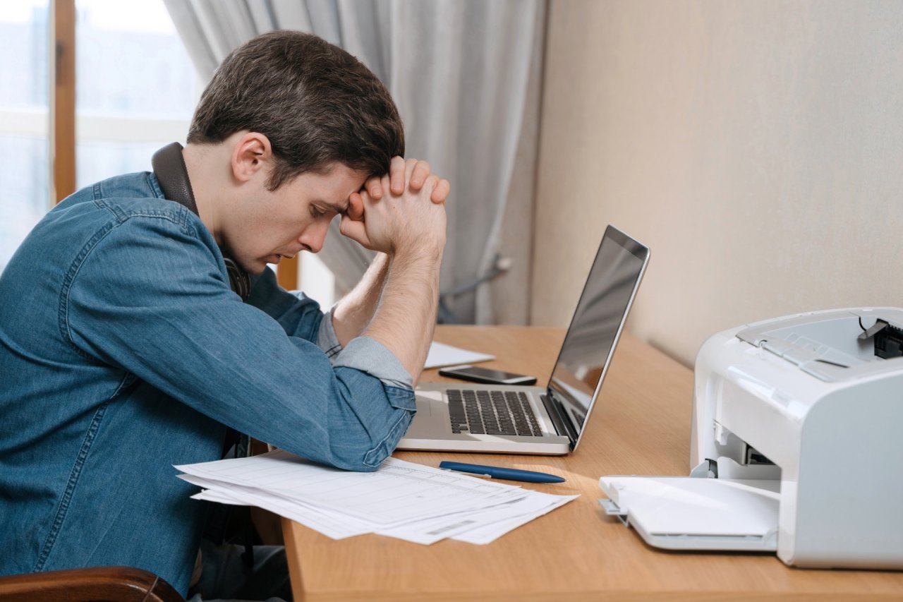 Youn man sat at an office desk looking stressed, head in his hands.