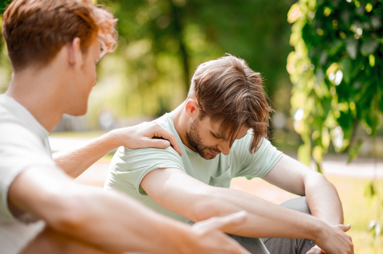 two men sat side by side on the floor, looking sad, one  with his hand on the others shoulder.