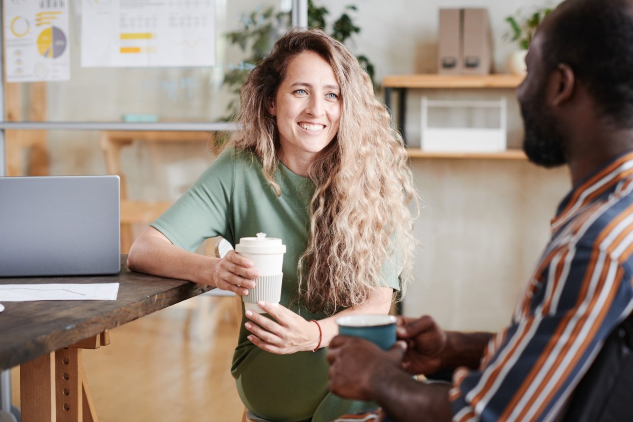 Blonde woman sat at a desk with a coffee, smiling and talking to a man.